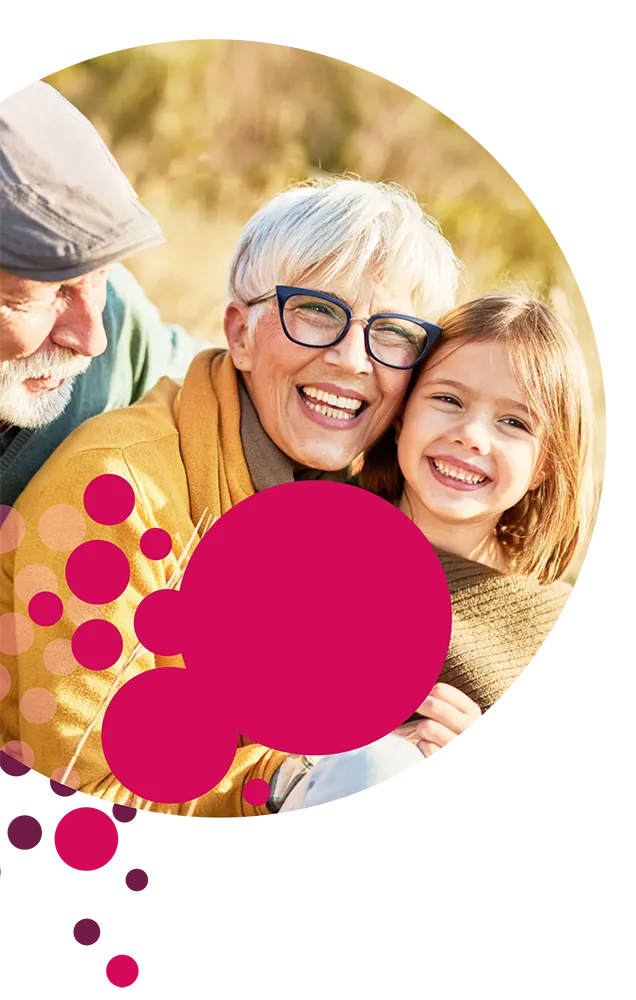 Älteres Paar und junges Mädchen lachen gemeinsam und umarmen sich draußen in warmer Herbstsonne. Die ältere Frau mit kurzen weißen Haaren und blauer Brille steht im Mittelpunkt, daneben ein lächelndes Kind. Links ist teilweise ein älterer Mann mit Mütze zu sehen. Über das Bild verteilt befinden sich dekorative pinke und violette Kreise.